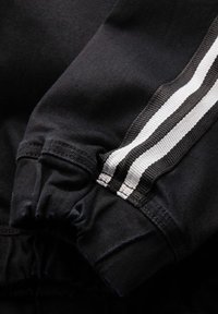 Jogger en tissu noir avec un accent rayé blanc le long du côté, doté d'un détail de poignet froncé à la cheville et d'une texture lisse.