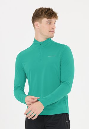 Mand i en lys grøn langærmet sportstrøje med "Endurance"-logo, kigger til siden, hænderne rører hinanden, hvid baggrund.