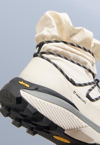Kremowy wysokotopowy but trekkingowy z teksturowanym materiałowym kołnierzem, czarnymi sznurowadłami i podeszwą Vibram z grubym bieżnikiem dla lepszej przyczepności. Widoczne logo Primaloft.