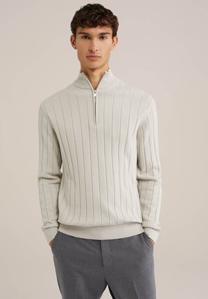 Suéter de cremallera gris claro con textura acanalada, presenta un cuello alto y puños ajustados. Llevado sobre pantalones gris oscuro.