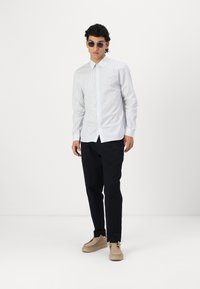 Camisa abotonada a rayas blancas y azules, pantalones negros y zapatos beige. El modelo lleva gafas de sol redondas. Diseño simple y moderno con un corte ajustado.