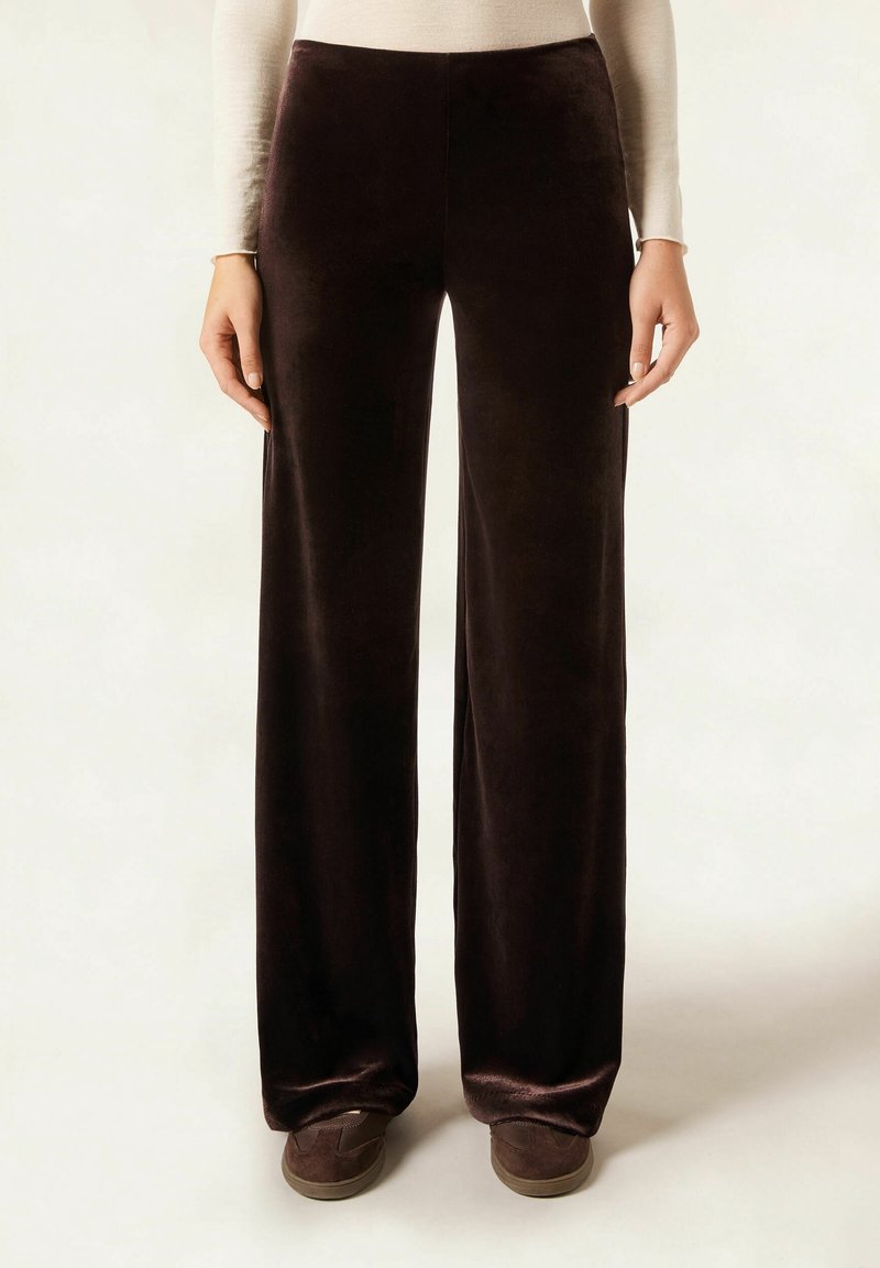 Femme portant un pantalon en velours marron foncé taille haute à jambes larges, associé à des chaussures marron foncé et un haut à manches longues beige.