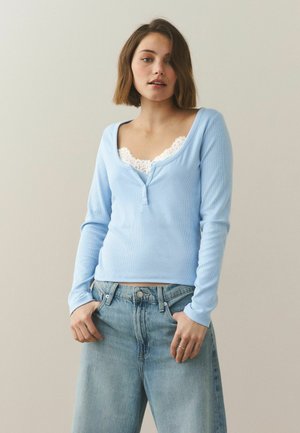 Ung kvinde iført en lyseblå langærmet top over en hvid camisole med blonder og løstsiddende lyseblå jeans, stående foran en ensfarvet baggrund.
