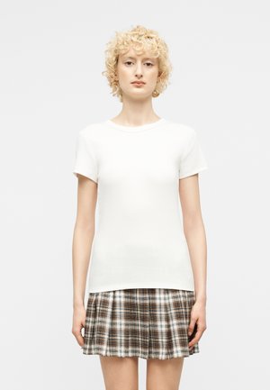 Rövid, göndör szőke hajú nő fehér rövid ujjú inget és barna-fehér kockás, pliszírozott szoknyát visel egyszínű háttér előtt.