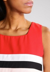 Červené šaty bez rukávů se čtvercovým výstřihem, s vodorovnými pruhy v bílé, černé a světlé růžové barvě na spodní části. Hladký povrch látky.