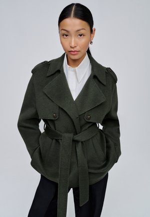 Femme aux cheveux foncés attachés, portant un manteau en laine vert foncé ceinturé par-dessus une chemise blanche et un pantalon noir, debout les mains dans les poches.