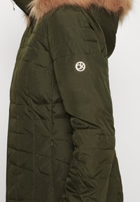 Veste matelassée verte avec texture capitonnée, dotée d'une capuche en fausse fourrure et d'un logo visible sur la manche. Détail zippé et design épuré.