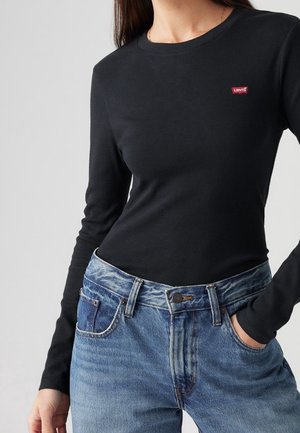 Femme portant un t-shirt noir à manches longues Levi's rentré dans un jean bleu taille haute, main dans la poche, sur un fond clair uni.
