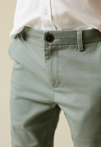 Pantaloni verde chiaro con una texture liscia, dotati di chiusura con bottone marrone e tasche frontali. Indossati con una camicia bianca con colletto.