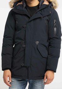 Marinblå parka med en huva fodrad med päls, quiltad textur, framfickor och en ficka med dragkedja på ärmen. Har svarta läderdetaljer.