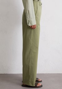 Green, high-waisted trousers made of lightweight fabric, featuring a straight fit, side pockets, and a clean hem, paired with dark sandals.