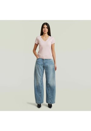 Vrouw staand met hand in zak, gekleed in lichtroze V-hals T-shirt, wijd uitlopende blauwe spijkerbroek en zwarte puntige schoenen, op een effen achtergrond.