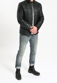 Homme portant une veste matelassée noire, un jean gris clair retroussé et des bottines noires, debout les bras croisés sur la taille.