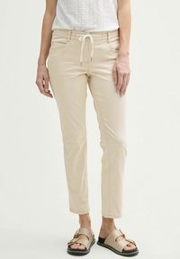 Pantaloni beige su misura con vita regolabile, dotati di tasche frontali e vestibilità slim. Abbinati a sandali dorati con fibbie.