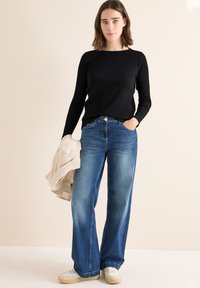 Zwarte geribbelde langemouwen top, gecombineerd met losse lichtblauwe jeans. Model houdt een crèmekleurige gestreepte jas vast en draagt witte sneakers.