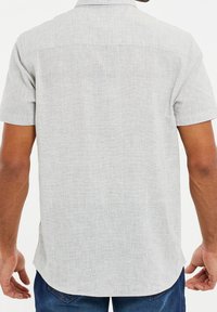 Camisa de manga corta en tejido gris claro con un patrón texturizado. Presenta un cuello abotonado y un dobladillo curvado, mostrada desde la parte de atrás.