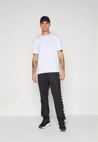 Tricou alb din bumbac cu mâneci scurte, asortat cu pantaloni sport negri cu dungi albe pe laterale. Pantofi sport negri și o șapcă completează ținuta.