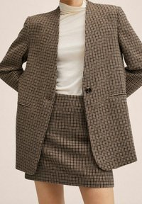 Blazer marrón a cuadros con solapas de muesca y cierre de botón único, combinado con una falda ajustada marrón a cuadros. Suéter de cuello vuelto blanco liso debajo.