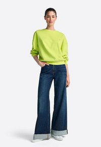 Élénksárga-zöld, lezser vállú pulóver, széles szárú, sötétkék farmerrel, felhajtott szegélyekkel és kontrasztos varrással párosítva.