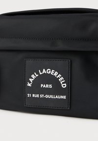 Melnā neilona soma ar taisnstūra formu, ar rāvējslēdzēju aizdari un teksturētu etiķeti ar baltu tekstu, kas norāda zīmolu un adresi.