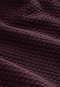 Texturiertes, tiefburgunderfarbenes Gewebe mit einem Diamantmuster, weiche Strickkonstruktion und ein leicht drapierter Look, der seine Tiefe und Textur zur Geltung bringt.