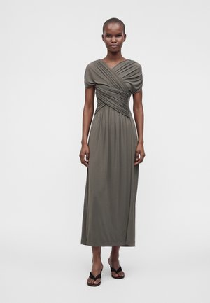 Žena v olivovozelených zavinovacích šatách s krátkym rukávom s plisovanými detailmi a čiernych otvorených sandáloch na podpätku, stojaca pred jednofarebným pozadím.
