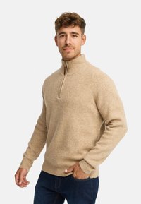 Pullover en tricot beige avec un col zippé mi-hauteur, présentant une texture côtelée et des manches longues. Associé à un jean bleu foncé.