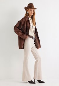 Bruine suède fringejack over een crème top, beige flarebroek en bruine puntige hakken. Grote zilveren riemgesp en blauwe oorbellen.