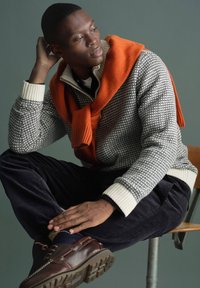 Man sitter på stol med handen på huvudet, bär en mönstrad tröja, orange halsduk över axlarna, svarta byxor och bruna skor.