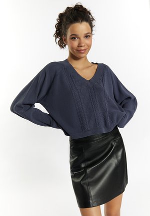 Marineblauer Kabelstrickpullover mit V-Ausschnitt und grob strukturiertem Strick, kombiniert mit einem eng anliegenden schwarzen Lederminirock.