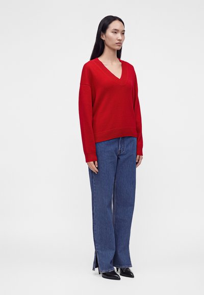 Femme aux longs cheveux noirs portant un pull rouge à col en V, un jean bleu à jambes larges avec des fentes latérales et des chaussures pointues noires, debout sur un fond blanc.