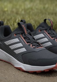Chaussures de sport gris foncé avec des bandes blanches, des accents orange, un marquage "TERREX" et des semelles texturées, posées sur un sol extérieur près de l'herbe.
