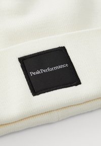 Cappello beanie crema realizzato in tessuto lavorato a maglia, con una toppa quadrata nera con "PeakPerformance" in testo bianco. Superficie testurizzata, bordo ripiegato.