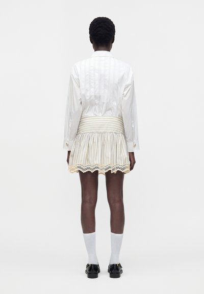 Personne debout de dos, portant une chemise blanche à manches longues, une jupe rayée avec bordure en dentelle, des chaussettes blanches et des chaussures noires sur un fond uni.