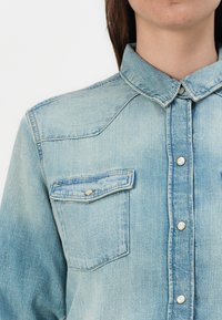 Camisa de denim azul claro com colarinho pontudo, botões de pressão em perolado e dois bolsos no peito com abas, apresentando um acabamento texturizado subtil.