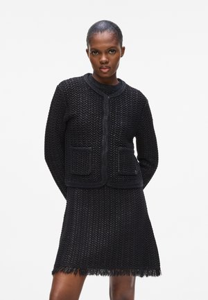 Veste en tricot noire avec un col rond et deux poches à l'avant, assortie d'une jupe à ourlet frangé. Tissu texturé dans l'ensemble.
