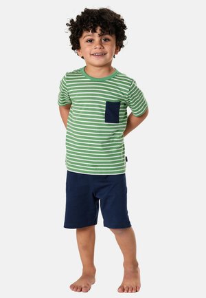 Young boy with curly hair smiling, wearing green and white striped t-shirt with blue pocket and navy blue shorts, standing barefoot.
