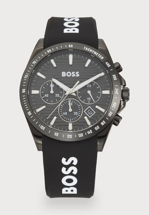 Black chronograph watch with a textured black dial, silver accents, and a rubber strap featuring white "BOSS" lettering. Date display at 6 o'clock.
