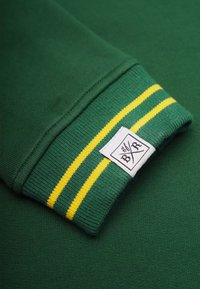 Grön sweatshirtärm med gula ränder, med en vit etikett sydd på. Tyget verkar slätt och elastiskt med en ribbad mudd.