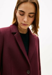Bordeauxwollen jas met een reverskraag, sluiting met één knoop en een gestructureerd zwart detail van een binnenvest. Glad materiaal, getailleerde pasvorm.