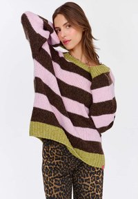Przesadzony sweter z wyrazistymi brązowymi i różowymi paskami, z zielonym ściągaczem. Noszony z spodniami w panterkę, ukazującymi dzianinową teksturę.
