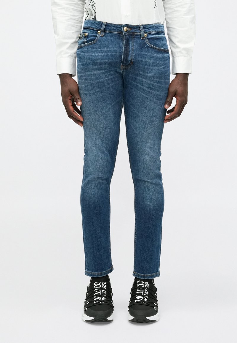 Vīrietis, kurš valkā šaurus zilo džinsu bikses, baltu garām piedurknēm kreklu un melnas kedas ar baltu tekstu uz vienkārša baltā fona.