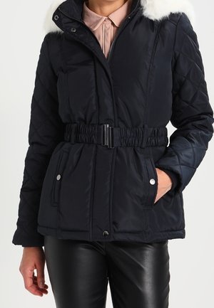 Frau trägt eine schwarze gesteppte Jacke mit tailliertem Gürtel, pelzbesetzter Kapuze und Taschen mit Druckknöpfen, kombiniert mit schwarzen Lederhosen.