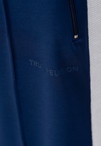 Pantalons de sport bleu marine en tissu texturé, avec un panneau latéral blanc et un logo en relief "TRUE RELIGION" sur le devant.