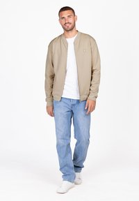 Chaqueta bomber con cremallera de color beige claro, con puños acanalados, llevada sobre una camiseta blanca, combinada con jeans azul claro y zapatillas blancas.