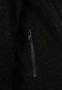 Schwarzer Fleece-Stoff mit einer strukturierten Oberfläche, ausgestattet mit einem seitlichen Reißverschluss aus Metall, der nahtlos in das Material integriert ist.