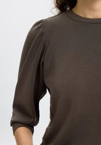 Maglietta marrone a maniche lunghe realizzata in un tessuto morbido, con spalle aderenti e maniche leggermente a sbuffo ai polsi. Design del collo semplice.