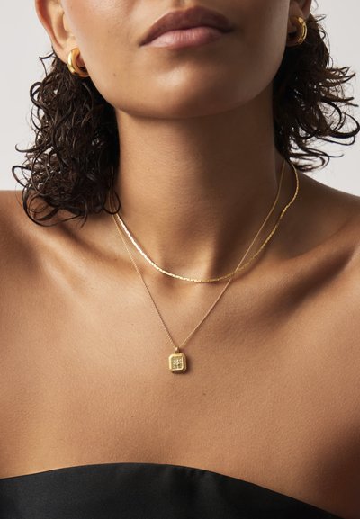 Οι χρυσές αλυσίδες με στρώση χρυσού διαθέτουν ένα υφασμένο τετράγωνο μενταγιόν και μία λεπτή αλυσίδα. Το μοντέλο φορά σκουλαρίκια κρίκους, με φόντο ένα σκούρο ρούχο.