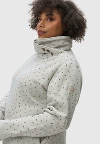 Light gray sweatshirt with a high collar, featuring a pattern of small triangles in black and red. Accented with two buttons and a logo patch.