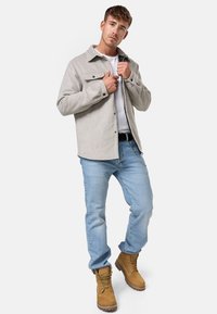 Camicia grigia con bottoni, maglietta bianca sotto, jeans blu e stivali da lavoro marroni. La camicia ha due tasche sul petto e una vestibilità comoda.